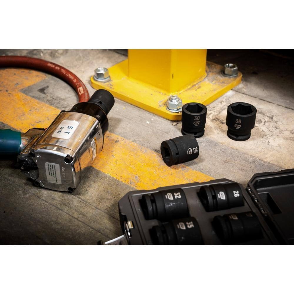 A professional shop floor scene showing the GEARWRENCH 3/4 inch drive heavy-duty metric impact sockets scattered next to a large pneumatic impact wrench and a yellow steel structural beam, ready for heavy equipment maintenance.