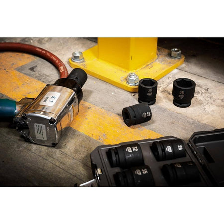 A professional shop floor scene showing the GEARWRENCH 3/4 inch drive heavy-duty metric impact sockets scattered next to a large pneumatic impact wrench and a yellow steel structural beam, ready for heavy equipment maintenance.