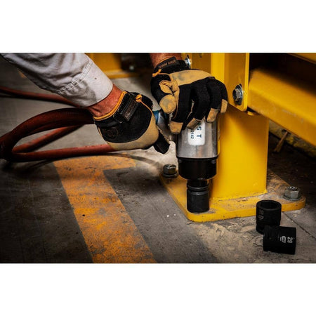 A professional mechanic wearing heavy work gloves uses a pneumatic impact wrench equipped with a GEARWRENCH 3/4 inch drive 6-point metric impact socket to secure a massive bolt on the yellow base of an industrial steel structure, with extra heavy-duty sockets resting on the concrete floor nearby.