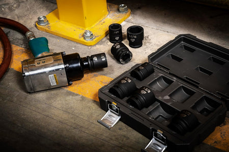 A lifestyle shot featuring a pneumatic impact wrench, the GEARWRENCH 84034 storage case, and several 3/4-inch drive SAE impact sockets scattered on a concrete floor. The image highlights the tool's use in industrial maintenance and professional repair settings.
