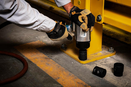 An action shot of a professional technician using a GEARWRENCH 3/4-inch drive impact socket with a pneumatic impact wrench. The worker is wearing protective gloves while securing a large heavy-duty bolt on a yellow industrial machine, demonstrating the tool's high-torque performance.