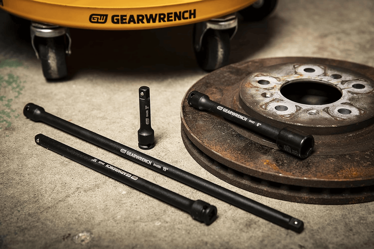 A collection of GEARWRENCH 3/8" drive impact extensions (3", 6", 10", 15") and a universal joint, arranged on a concrete floor next to a rusty brake rotor and a yellow GEARWRENCH tool cart base.