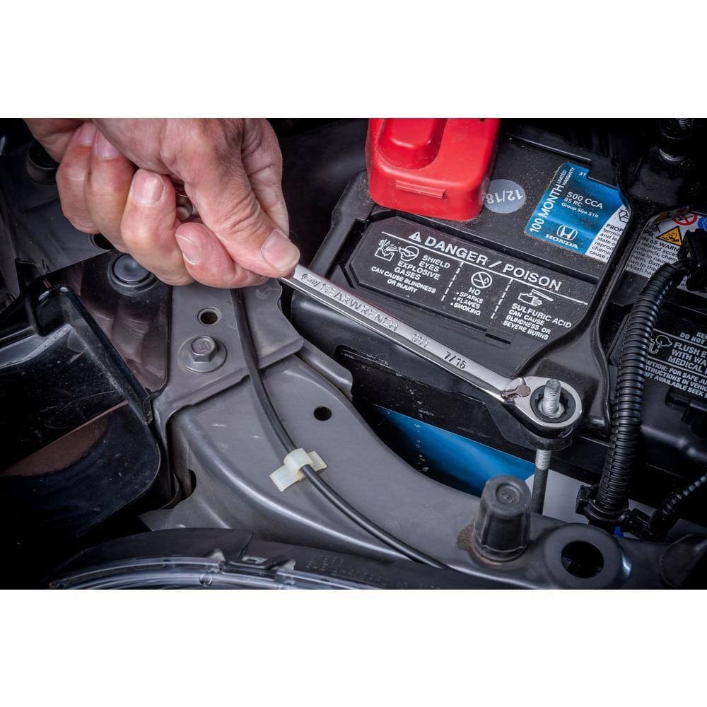 A close-up action shot of the GEARWRENCH 7/16 inch reversible ratcheting wrench being used to remove a bolt near a vehicle battery, highlighting the efficiency of the flush-mounted reversing lever in tight engine bays.