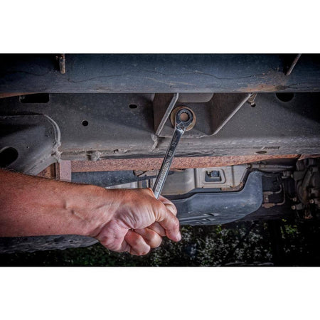 An action shot from beneath a vehicle showing a technician using the GEARWRENCH 1/2 inch combination wrench to access a chassis bolt, demonstrating the tool's durability in rugged automotive environments.