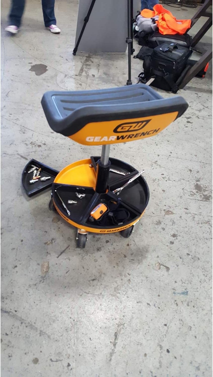 A perspective shot of the GEARWRENCH adjustable mechanics seat in use, highlighting the utility of the magnetic base trays for holding specialized automotive tools like headlamps and fasteners.