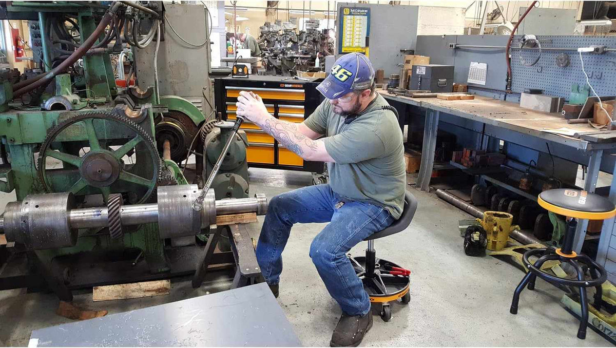 A professional technician using the GEARWRENCH 86994 rolling mechanics seat while working on a large industrial machine, demonstrating the stool's stability and ergonomic support during heavy-duty maintenance tasks.