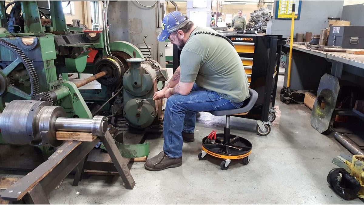 An action photo showing a mechanic seated on the GEARWRENCH rolling stool while performing repairs, with tools and parts organized in the integrated magnetic trays for high-efficiency workflow.