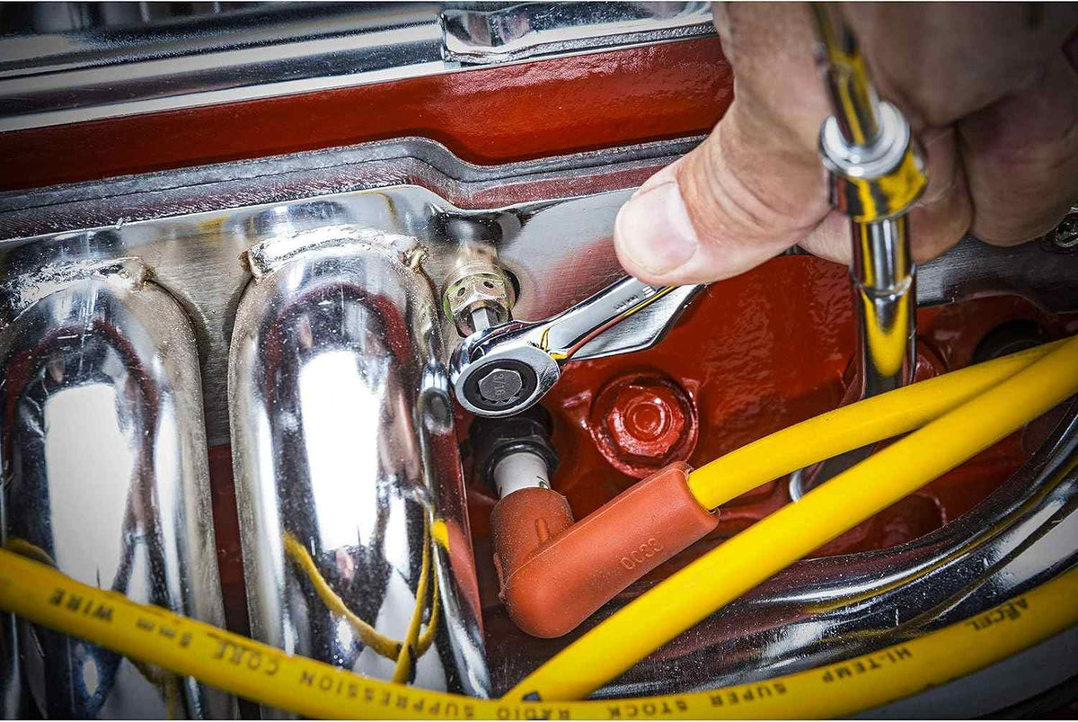 A hand uses a small, flex-head ratcheting wrench to access a bolt in a tight engine bay, next to a red valve cover and yellow spark plug wires.