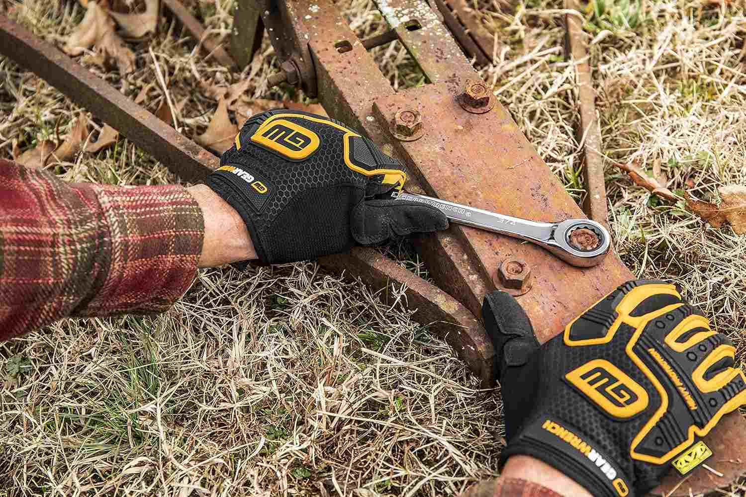 A person's gloved hands use a large GEARWRENCH ratcheting combination wrench on a rusty bolt on a piece of outdoor farm equipment.
