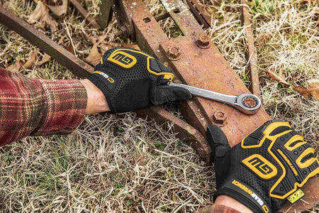A person's gloved hands use a large GEARWRENCH ratcheting combination wrench on a rusty bolt on a piece of outdoor farm equipment.