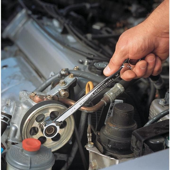 An action photo of a technician using a GEARWRENCH reversible ratcheting wrench to perform maintenance on a vehicle's engine pulley, demonstrating the tool's accessibility in cramped automotive environments.