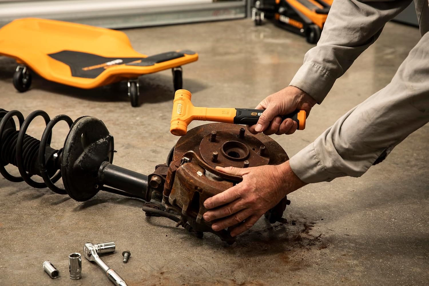 An action-shot view of an automotive service professional in a garage environment using a handheld orange GEARWRENCH dead blow hammer to break free a rusted vehicle brake rotor and wheel hub assembly.