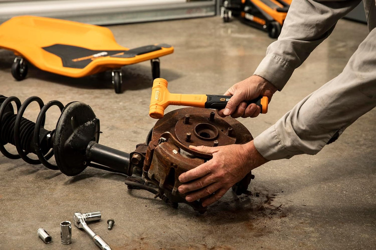 An action-shot perspective of an automotive technician in a commercial repair garage using a standard orange GEARWRENCH dead blow hammer to break free and service a rusted vehicle wheel hub and brake rotor assembly.