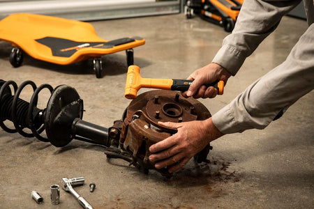 An action-shot perspective of an automotive technician in a commercial repair garage using a standard orange GEARWRENCH dead blow hammer to break free and service a rusted vehicle wheel hub and brake rotor assembly.