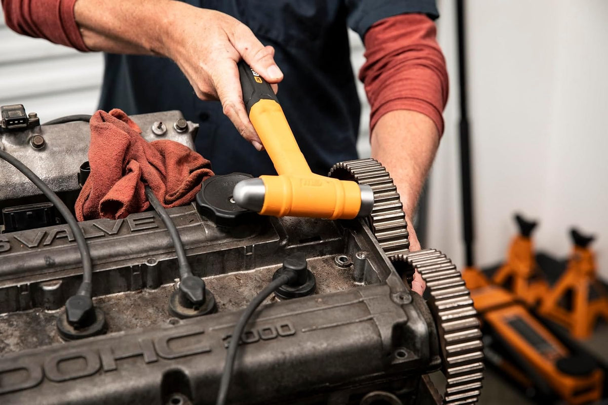 An in-use detail shot showing an auto service professional using a GEARWRENCH ball pein dead blow hammer to align a timing gear on a double overhead cam (DOHC) vehicle engine block during a drivetrain cylinder head repair.