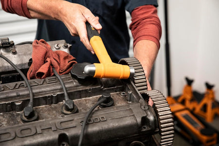 An in-use detail shot showing an auto service professional using a GEARWRENCH ball pein dead blow hammer to align a timing gear on a double overhead cam (DOHC) vehicle engine block during a drivetrain cylinder head repair.