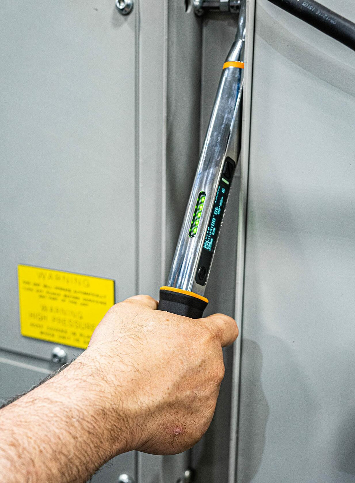 A worker using the GEARWRENCH electronic torque wrench to tighten a fastener in a confined vertical space on a metal enclosure, with the digital display visible.