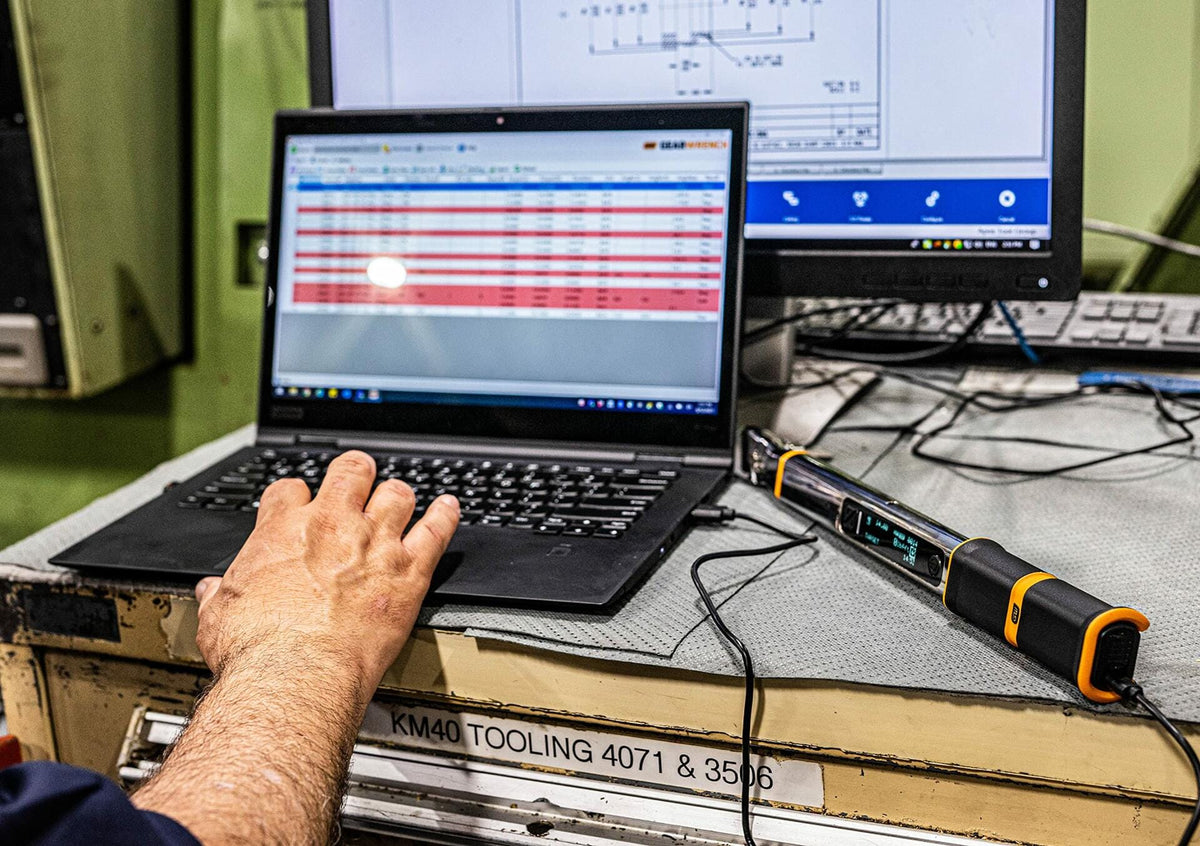 The GEARWRENCH electronic torque wrench connected to a laptop computer via USB cable, with a hand on the keyboard, demonstrating its data download and connectivity feature in a shop environment.