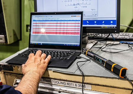 The GEARWRENCH electronic torque wrench connected to a laptop computer via USB cable, with a hand on the keyboard, demonstrating its data download and connectivity feature in a shop environment.