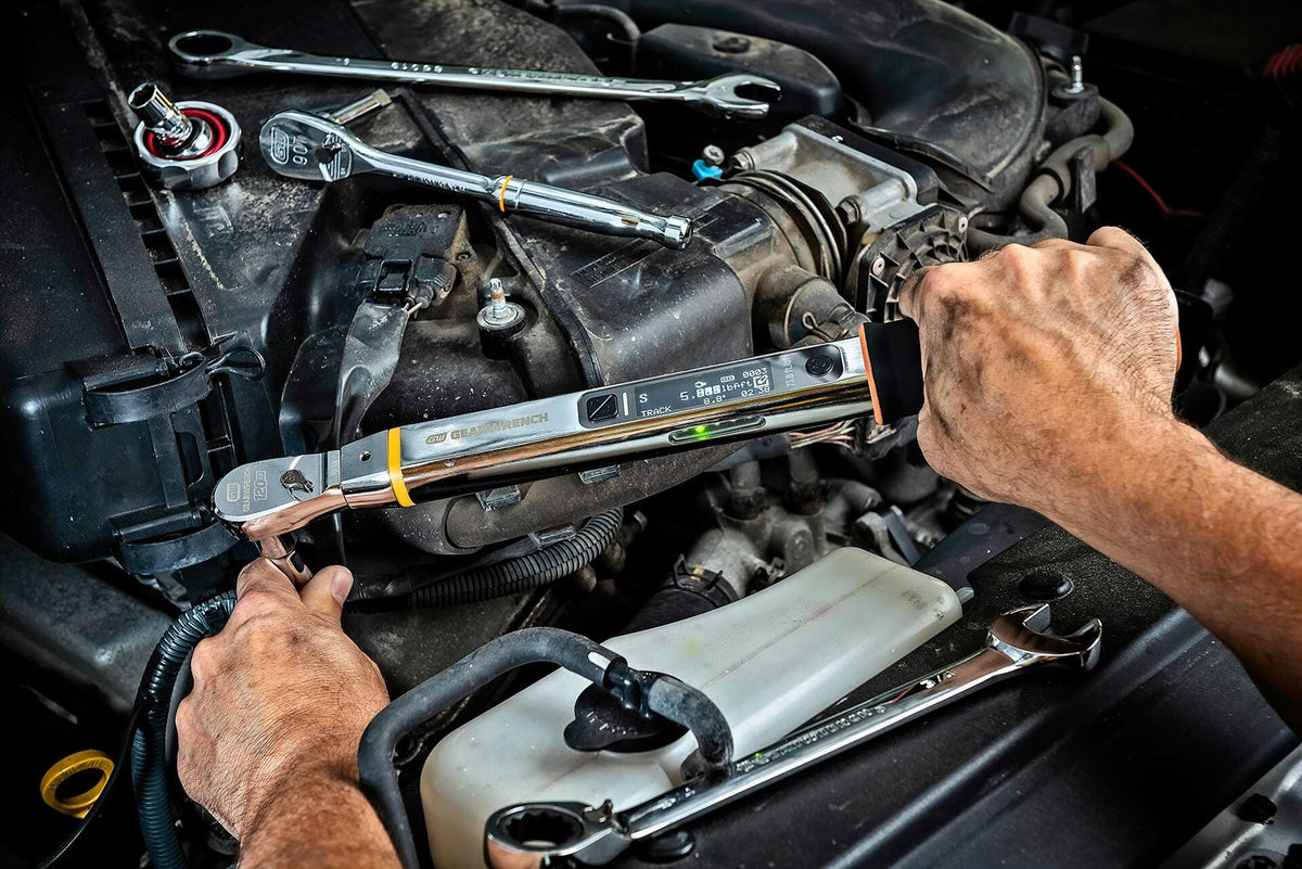 A mechanic's hands using the GEARWRENCH electronic torque wrench to fasten a bolt in a vehicle engine bay, with other chrome wrenches resting nearby.