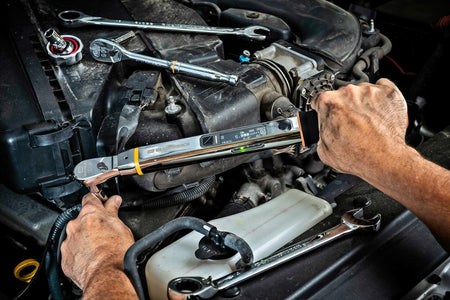 A mechanic's hands using the GEARWRENCH electronic torque wrench to fasten a bolt in a vehicle engine bay, with other chrome wrenches resting nearby.