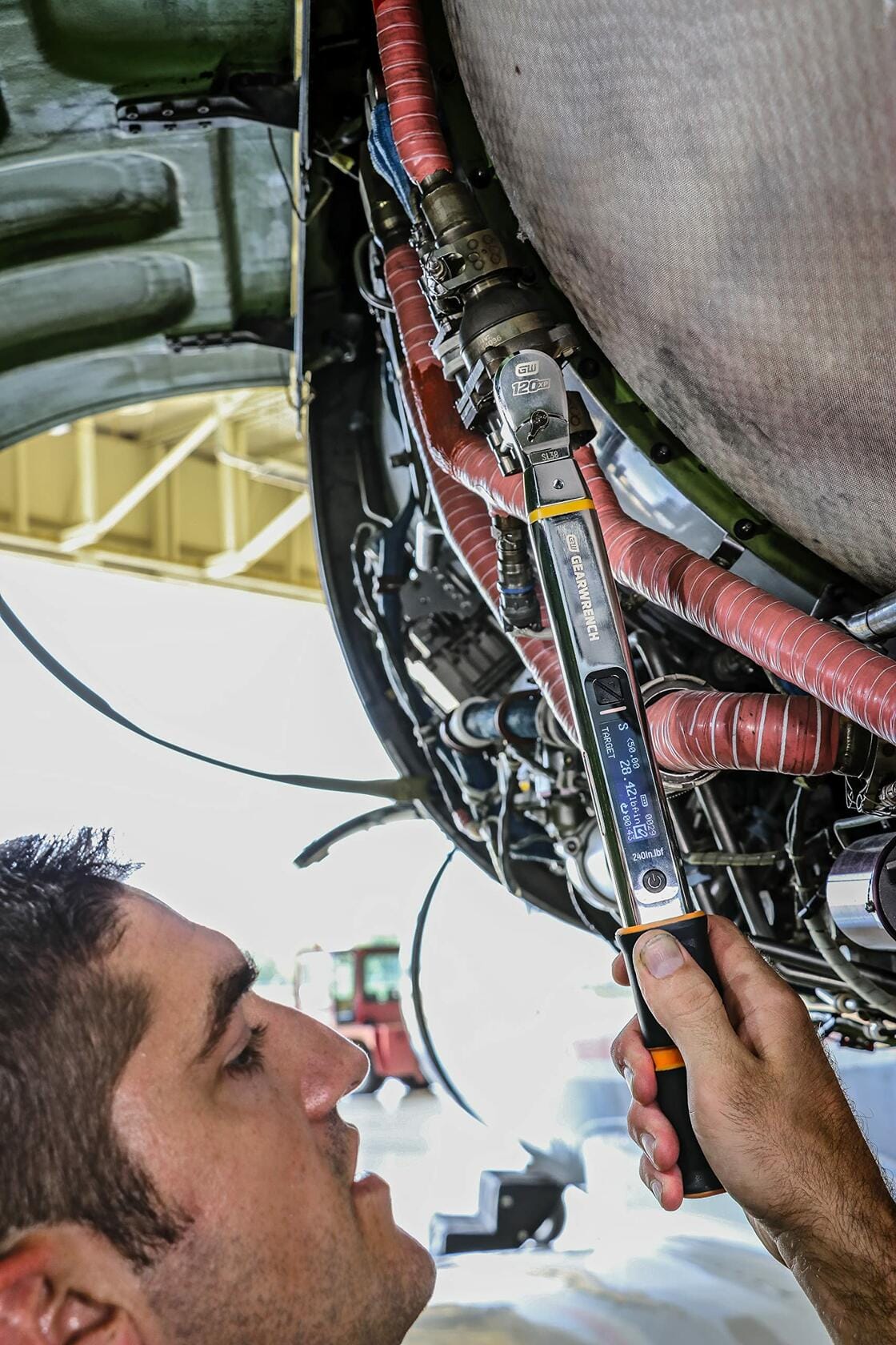 A technician working overhead on an intricate machine or engine, using the GEARWRENCH electronic torque wrench to apply precise torque and angle to a fastener in a confined space.