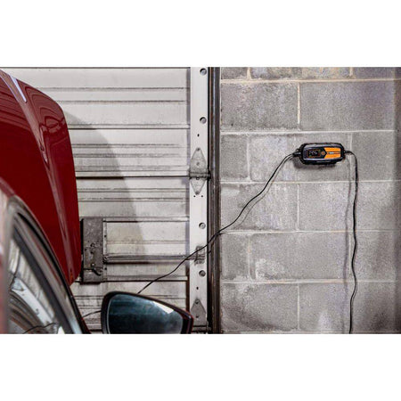 A lifestyle shot showing the GEARWRENCH battery maintainer mounted to a gray cinderblock garage wall. The unit is connected to a vehicle, demonstrating the long reach of the power cords and the convenience of the wall-mountable design for long-term vehicle storage.