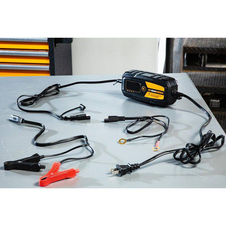 A complete product kit view of the GEARWRENCH GWBC015 battery charger and maintainer set laid out on a workshop table. The image shows the 1.5A smart charger unit, the AC power cord, heavy-duty battery clamps with quick-disconnect plugs, and the permanent ring terminal leads for long-term vehicle battery conditioning.