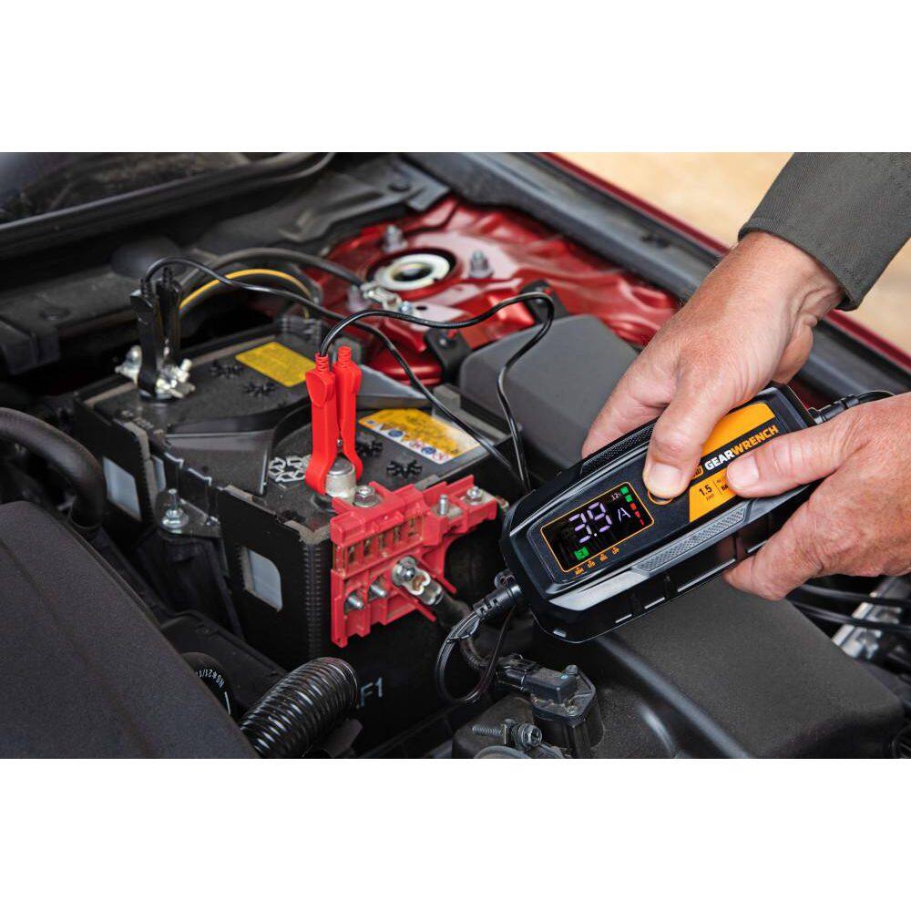 An action lifestyle shot of a professional using the GEARWRENCH maintainer on a modern car battery. The worker's hand is visible holding the unit while the red and black clamps are secured to the battery posts, with the digital display showing a live 3.9 Amp charging current.