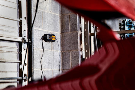 A lifestyle shot showing the GEARWRENCH GWBC030 smart charger mounted to a gray cinderblock garage wall. The unit is connected to a vehicle, demonstrating the convenience of the wall-mountable design for professional workshops and long-term vehicle maintenance.