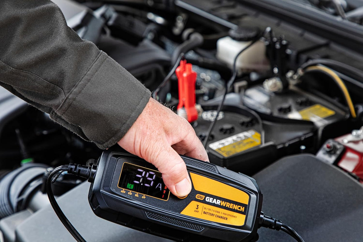 An action lifestyle shot of a technician using the GEARWRENCH 3A maintainer on a modern vehicle. The worker's hand is shown holding the unit while the digital display indicates a live 3.9 Amp charging status, demonstrating real-time battery diagnostics under the hood.