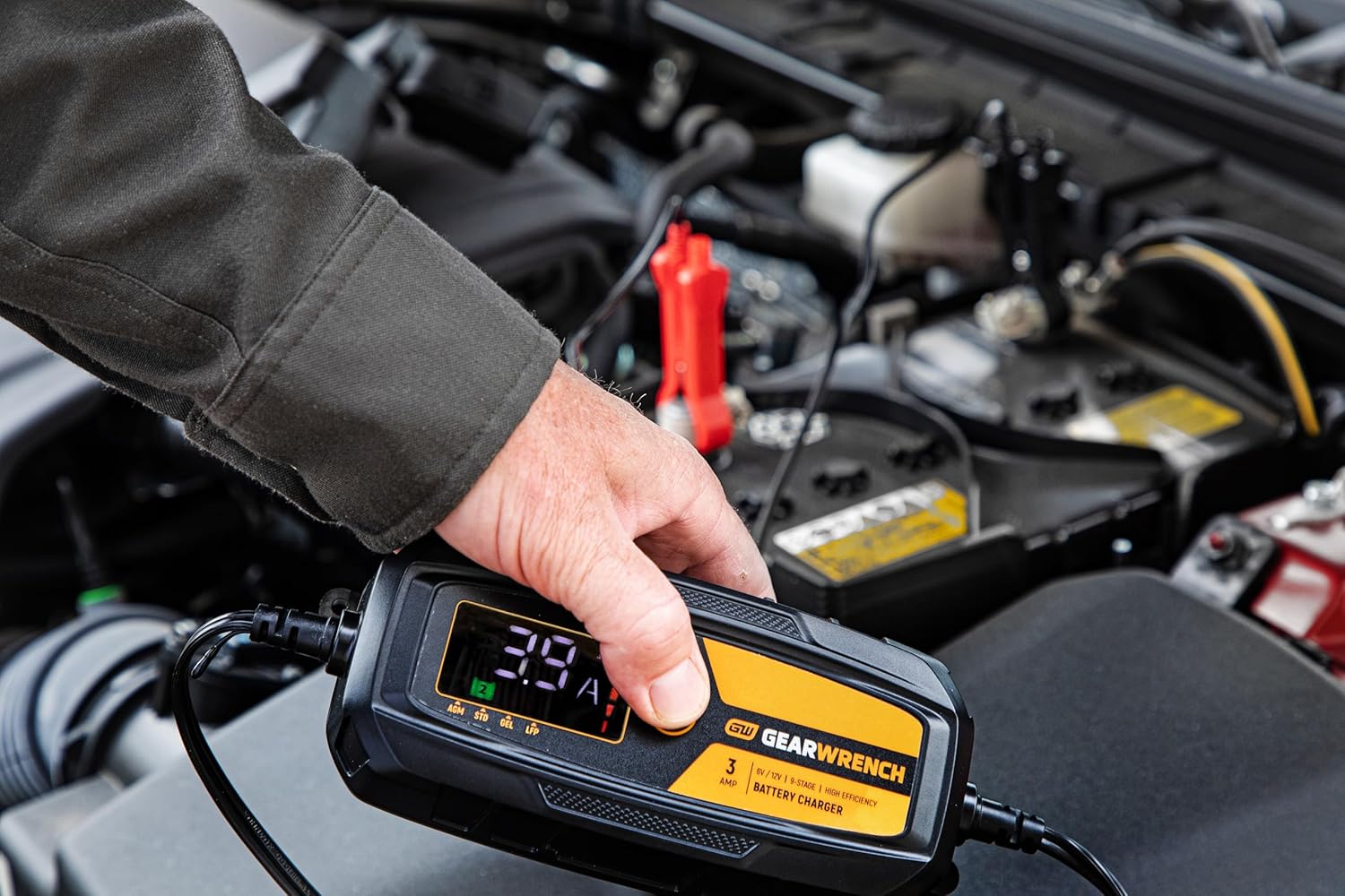 An action lifestyle shot of a technician using the GEARWRENCH 3A maintainer on a modern vehicle. The worker's hand is shown holding the unit while the digital display indicates a live 3.9 Amp charging status, demonstrating real-time battery diagnostics under the hood.