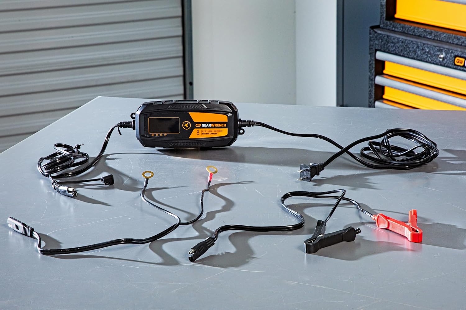A complete product kit view of the GEARWRENCH GWBC030 3A battery charger set laid out on a workshop surface. The image shows the smart charger unit, the AC power cord, heavy-duty battery clamps with quick-disconnect plugs, and the permanent ring terminal leads.