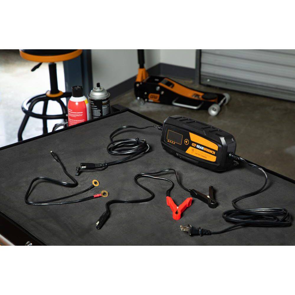 A complete product kit view of the GEARWRENCH GWBC100 10A battery charger set laid out on a workshop surface. The image shows the smart charger unit, the AC power cord, heavy-duty battery clamps with quick-disconnect plugs, and the permanent ring terminal leads.