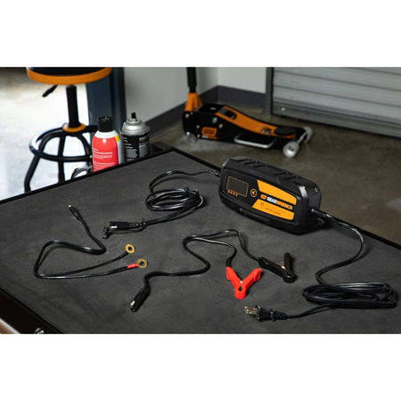 A complete product kit view of the GEARWRENCH GWBC100 10A battery charger set laid out on a workshop surface. The image shows the smart charger unit, the AC power cord, heavy-duty battery clamps with quick-disconnect plugs, and the permanent ring terminal leads.