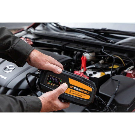 An action lifestyle shot of a technician using the GEARWRENCH 10A maintainer on a modern vehicle. The worker's hands are shown holding the unit while the digital display indicates a 14.4V charging status, demonstrating real-time battery diagnostics under the hood.