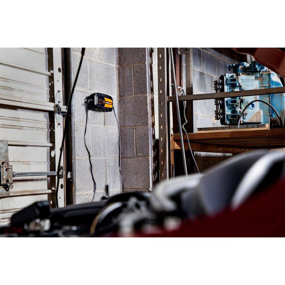 A lifestyle shot showing the GEARWRENCH GWBC100 smart charger mounted to a gray cinderblock garage wall. The unit is connected to a vehicle, demonstrating the convenience of the wall-mountable design for professional workshops and long-term vehicle maintenance.