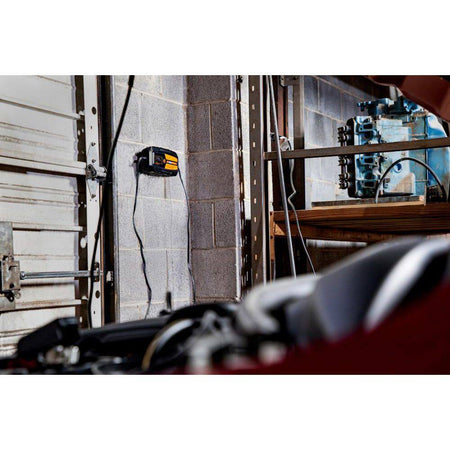 A lifestyle shot showing the GEARWRENCH GWBC100 smart charger mounted to a gray cinderblock garage wall. The unit is connected to a vehicle, demonstrating the convenience of the wall-mountable design for professional workshops and long-term vehicle maintenance.
