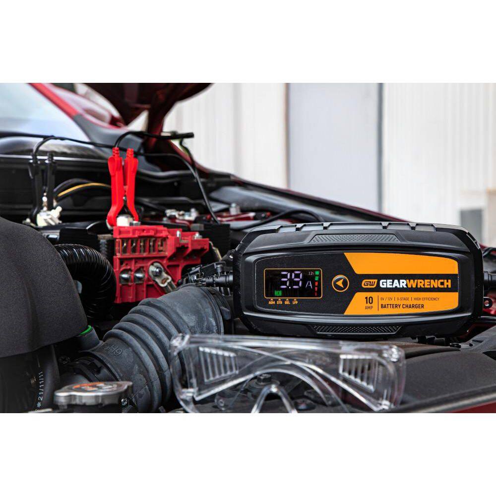 A wide lifestyle shot of the GWBC100 unit resting on a vehicle's engine shroud during a maintenance session. The bright molten orange housing is clearly visible against the engine components, demonstrating its professional utility and 10-Amp high-efficiency charging power.