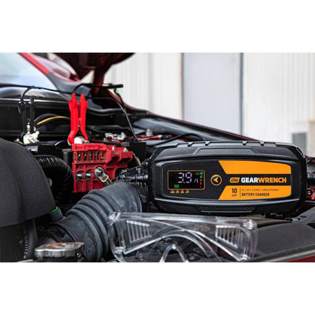 A wide lifestyle shot of the GWBC100 unit resting on a vehicle's engine shroud during a maintenance session. The bright molten orange housing is clearly visible against the engine components, demonstrating its professional utility and 10-Amp high-efficiency charging power.