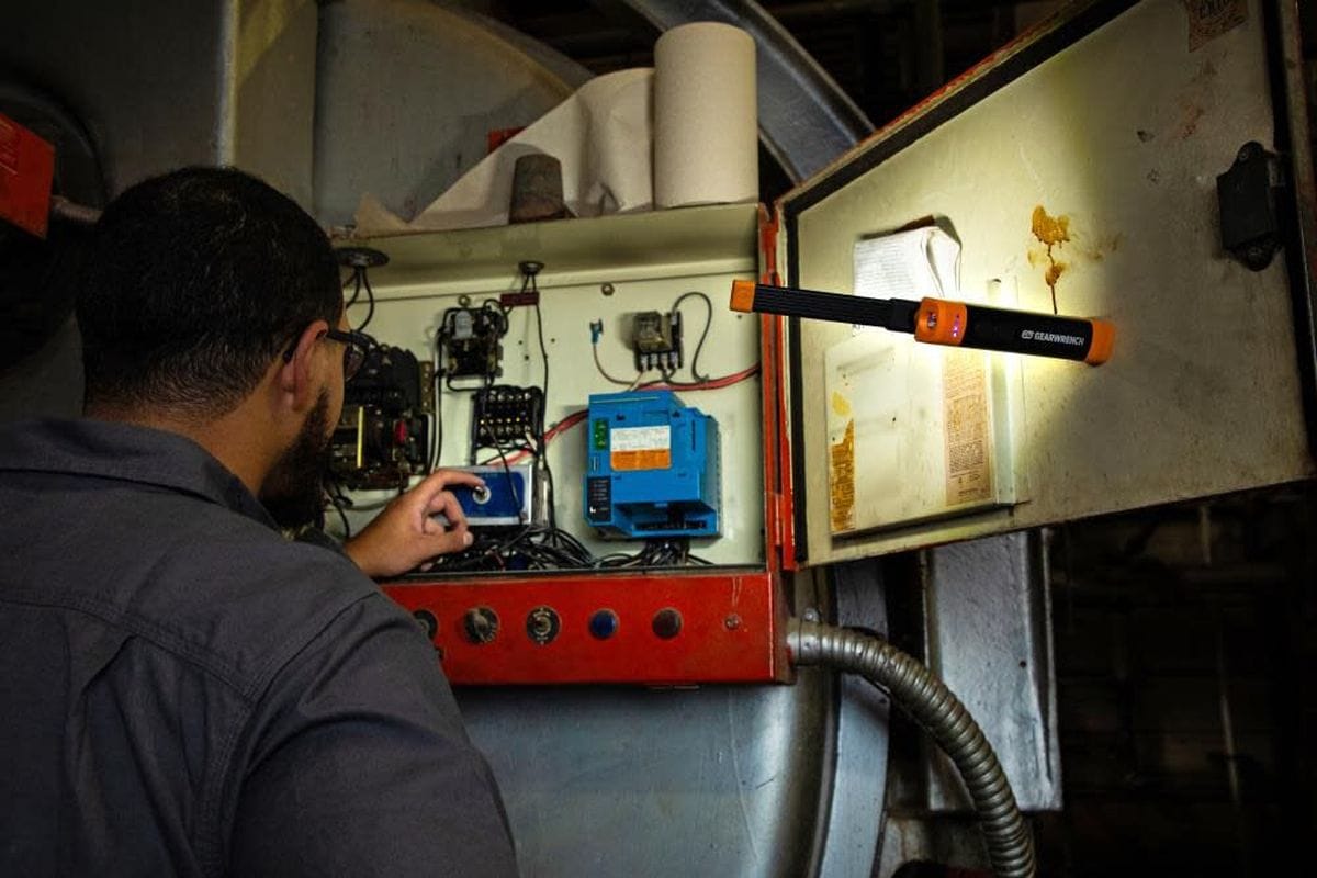 Action shot showing a professional mechanic using the magnetic base of the GEARWRENCH GWFL105 flex head light to illuminate a large industrial electrical control panel hands-free.
