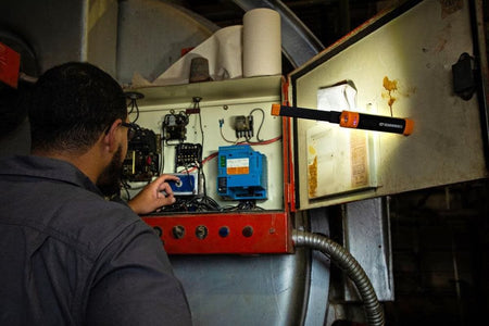 Action shot showing a professional mechanic using the magnetic base of the GEARWRENCH GWFL105 flex head light to illuminate a large industrial electrical control panel hands-free.