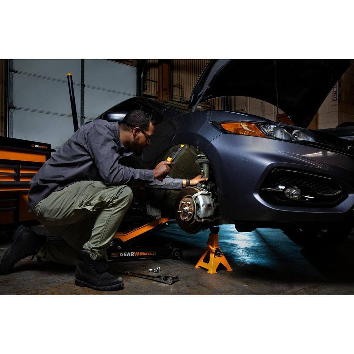 A technician using the GEARWRENCH GWFL105 rechargeable light to illuminate the wheel well and suspension components of a car while it is supported by a jack stand.