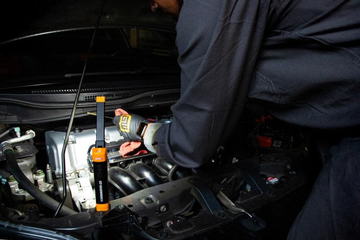 A professional technician using the magnetic base of the GEARWRENCH work light to attach it to a car hood while performing an engine inspection and oil level check.