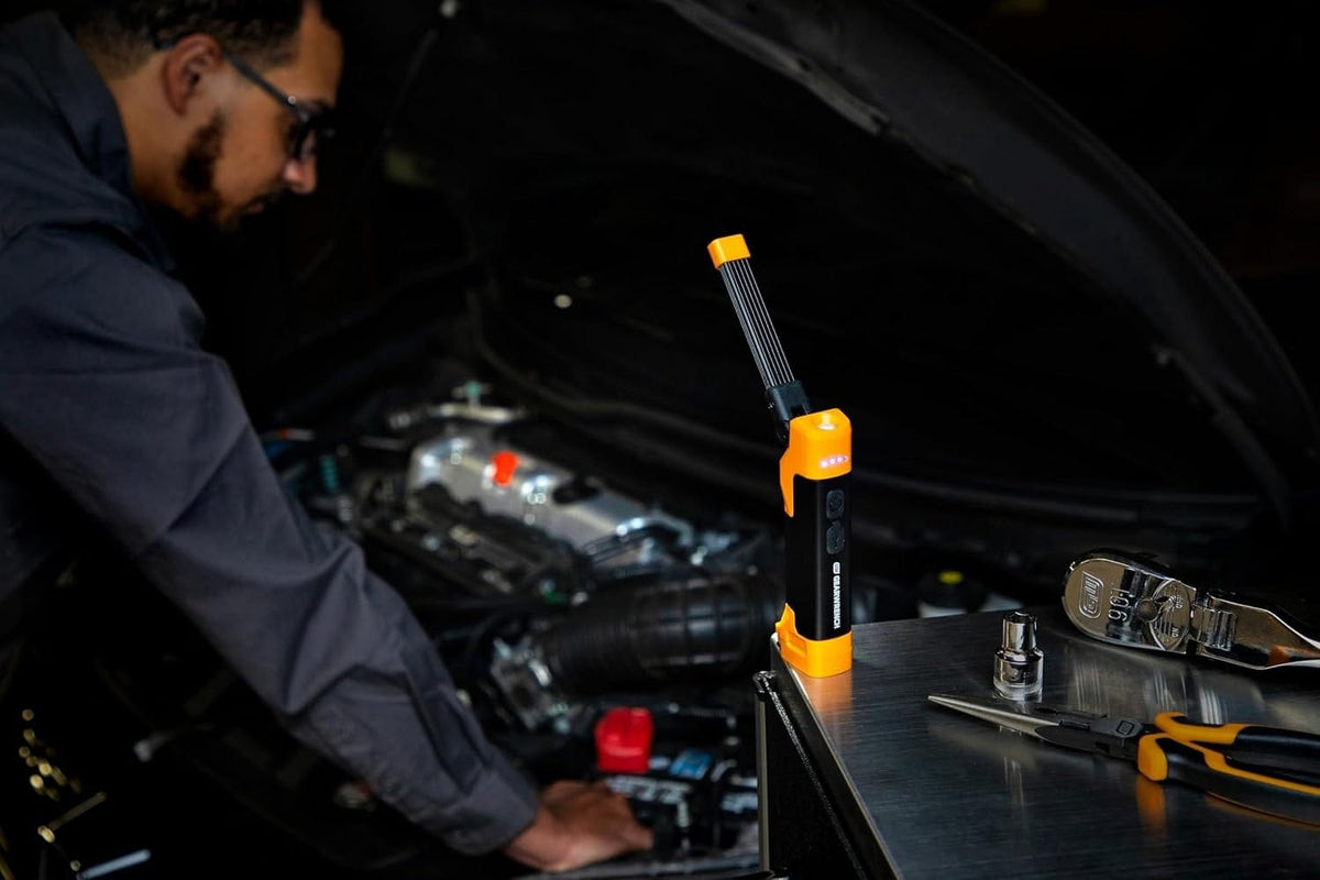 Action shot of a technician using the GEARWRENCH magnetic flex head LED work light to illuminate a dark engine bay during professional automotive maintenance, highlighting the light's 20-hour runtime.