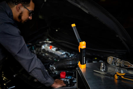 Action shot of a technician using the GEARWRENCH magnetic flex head LED work light to illuminate a dark engine bay during professional automotive maintenance, highlighting the light's 20-hour runtime.