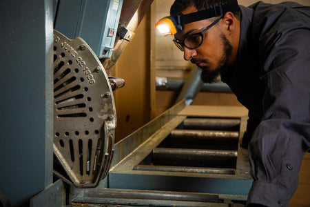 A worker wearing the lit GEARWRENCH headlamp bends to look closely at industrial conveyor rollers.