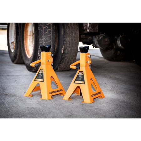 Two GEARWRENCH 12-ton heavy-duty jack stands positioned on a concrete shop floor next to a large truck tire, demonstrating professional load displacement and stability in an industrial setting.