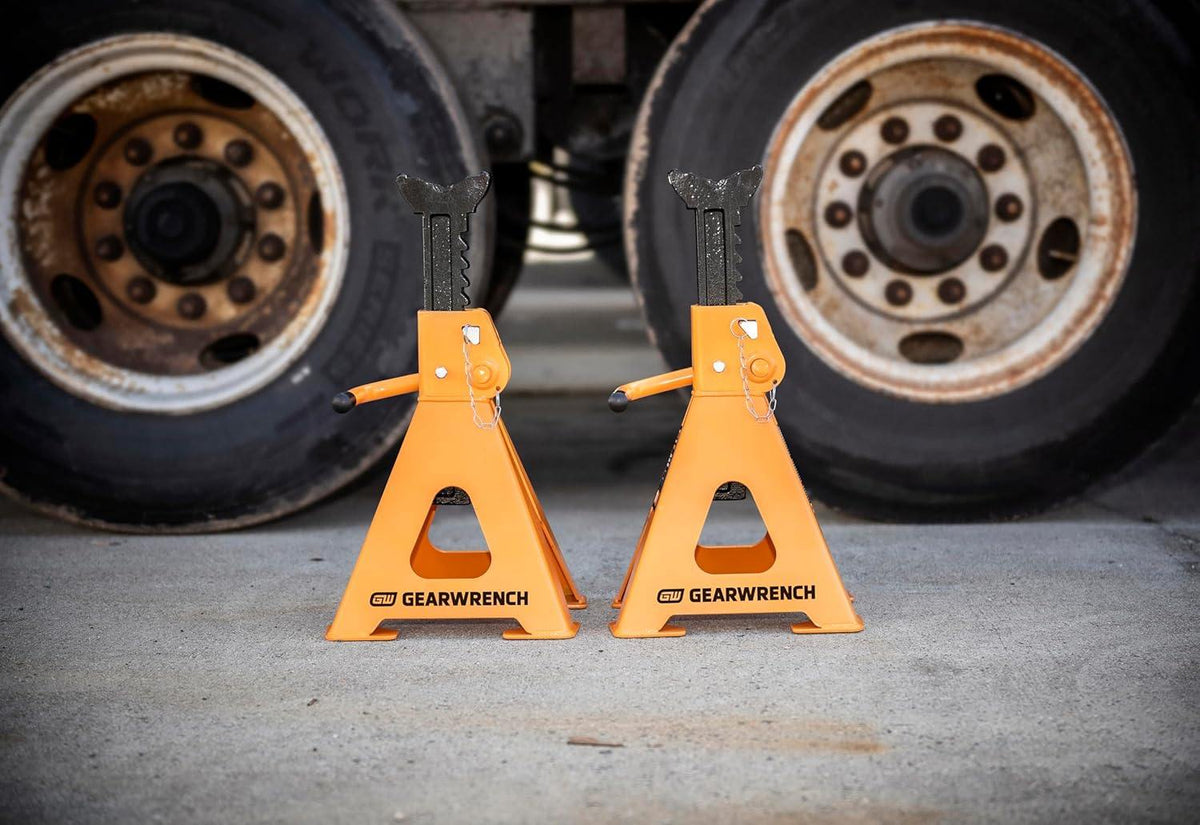Action shot of a pair of GEARWRENCH 12-ton molten orange jack stands supporting a heavy-duty vehicle axle, illustrating their high-capacity performance and ASME PASE standard compliance.