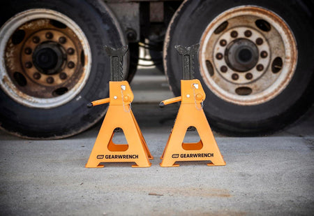 Action shot of a pair of GEARWRENCH 12-ton molten orange jack stands supporting a heavy-duty vehicle axle, illustrating their high-capacity performance and ASME PASE standard compliance.